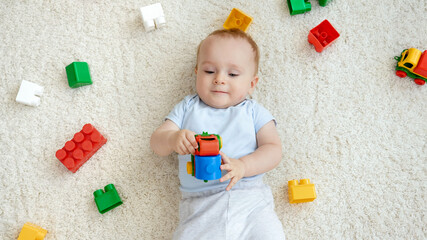 Top view of smiling baby boy lying on carpet in playroom with lots of colorful toys. Concept of children development, education and creativity at home