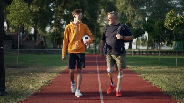 Front view wide shot happy motivated amputee sportsman and teenage boy walking on running track in summer park talking gesturing high-five. Caucasian father and son enjoying weekend sport outdoors