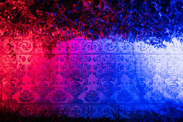Obraz premium Tiled wall partially covered with leaves, illuminated with red and blue neon lights. Traditional Portuguese tiles lit by a toxic light. Azulejo wall texture with vibrant pink and ultraviolet shine