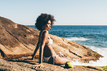 A ravishing young African American woman in white bikini and with curly afro hair is enjoying a sunset on the beach of a tropical resort while sitting on a rock in front of the ocean - obrazy, fototapety, plakaty