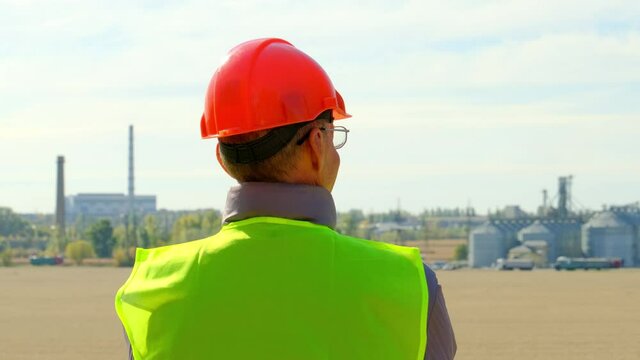 Grain elevator and silo tanks at farm. Professional builder in uniform at inspection checks powerful plant standing in field backside view