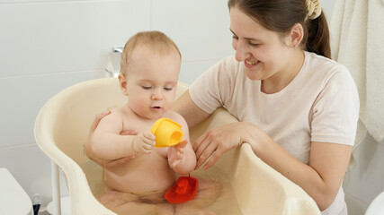 Happy smiling mother looking at her baby son washing in bathroom and playing toys