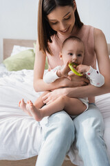 smiling woman holding baby boy with rattle ring while sitting on bed.