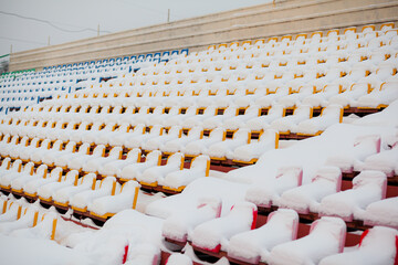 Empty outdoor football soccer stadium seats covered with snow in winter, light snowfall. Plastic seats in a row at the stadium are covered with snow snow.