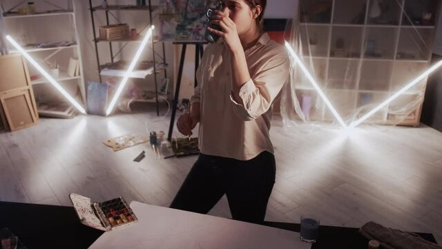 Female artist. Work pause. Creative process. Thoughtful woman drinking tea while drawing on map paper in dark light studio interior.