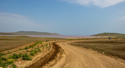 An empty dirt road near a pink lake on a summer day against the sky