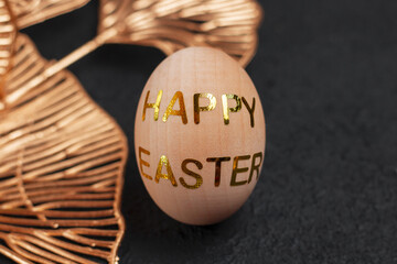 Wooden decorative egg with gold colored stickers with the inscription HAPPY EASTER. Concept for a postcard for Easter on a black textured background.