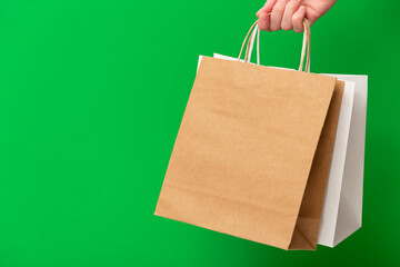 Female hand holding two shopping bags isolated on green background. White and brown craft blank paper bags in hand. Black friday sale, discount, shopping and ecology concept