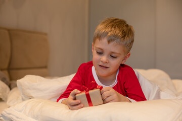 A little boy in Christmas pajamas unpacks a gift while lying in bed.