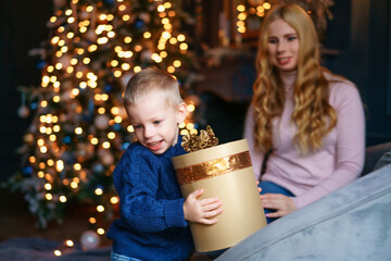 Fototapeta na wymiar Happy boy received large gift box from his mother, sitting on sofa by christmas tree. Christmas lights shine brightly on the tree. Christmas and new year family holiday concept