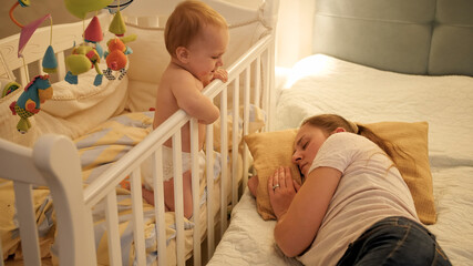 Young tired mother fell asleep next to her baby crying in bed. Concept of parenting, parent fatigue...