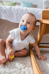 cheerful little boy with pacifier holding toy building block while sitting on floor.
