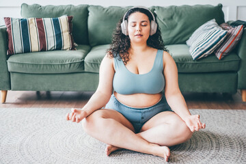 Curly haired plus size young woman in wireless headphones turns on music and meditates sitting in lotus position on floor mat against green sofa - obrazy, fototapety, plakaty