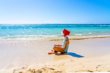 Fototapeta na wymiar Beautiful girl in a Christmas hat meditating on the tropical summer beach. Winter holiday on a tropical island concept.