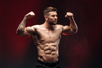 Obraz premium Young athletic man with a naked torso showing his biceps isolated on a red background. Bodybuilding and strong people concept