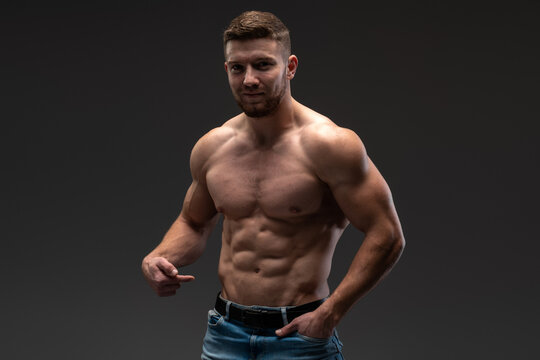 Waist up portrait of bearded shirtless bodybuilder looking at the camera while posing over the black wall. Stock photo