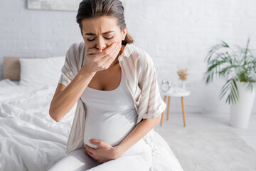 young pregnant woman feeling nausea and covering mouth in bedroom.
