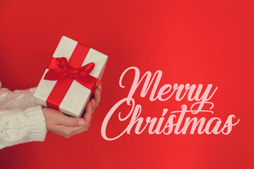 Fototapeta na wymiar Merry christmas text. Female wearing white knitted sweater holding a presents over isolated red background. Woman with a wrapped gift. Close up, copy space for text.