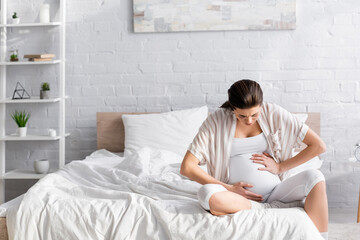 young pregnant woman suffering from cramp while sitting on bed.