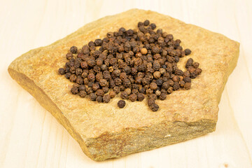 Lot of whole spicy black pepper with natural flat rock on plain wood