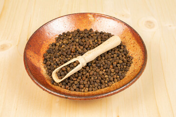 Lot of whole spicy black pepper in a wooden scoop in ceramic bowl on plain wood