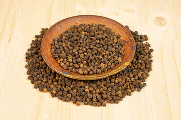 Lot of whole spicy black pepper in ceramic bowl on plain wood with pepper placed around