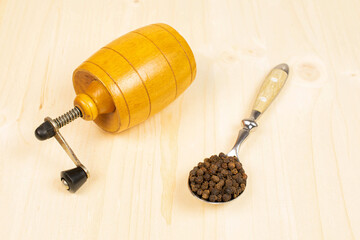 Lot of whole spicy black pepper with metal spoon with pepper mill on plain wood