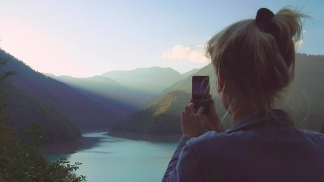 A slender, beautiful blonde takes photos or videos of a landscape in the autumn mountains. Georgia. A traveler in a denim suit. She looks at her smartphone, social networks.