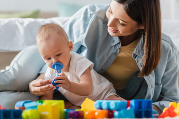 Obraz premium smiling woman with baby boy playing with blurred building blocks on floor.