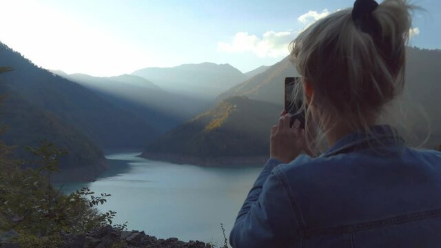 A slender, beautiful blonde takes photos or videos of a landscape in the autumn mountains. Georgia. A traveler in a denim suit. She looks at her smartphone, social networks.