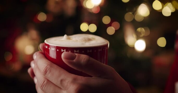 Close-up of women's hands holding a mug with hot drink beverage in Christmas entourage against the background of lights and a Christmas tree.