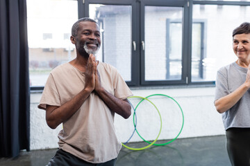 Smiling african american man practicing yoga near senior friend in sports center.