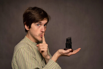 Young handsome tall slim white man with brown hair holding black crystal having finger on mouth in striped shirt on grey background