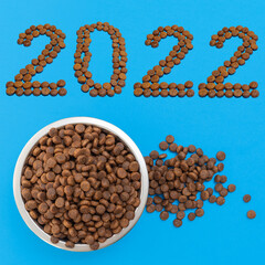 Date 2022 composed of dry food and a round metal bowl for dogs or cats full of crunchy dry food on a blue background
