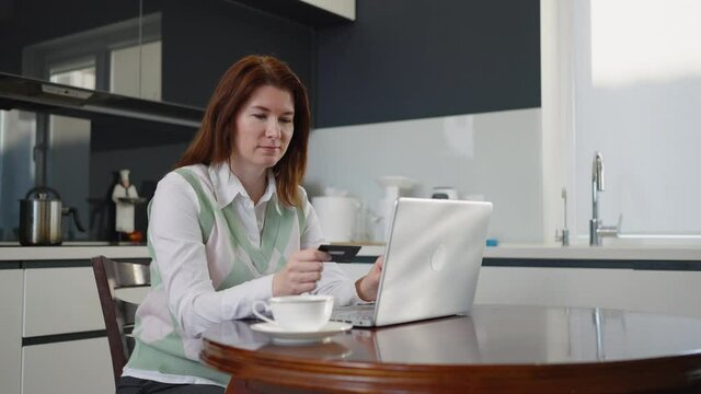 Adult woman making online order sitting in kitchen, holding credit card and entering numbers on laptop, buying what she wanted and making yes gesture with arm. Concept of shopping
