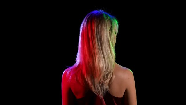 Back view of a young pretty blonde flirtatiously waving her hair. Girl model posing on black studio background with neon lights. Close up. Slow motion.
