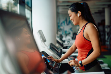 Happy Asian athlete adjusts speed on treadmill during sports training at gym.