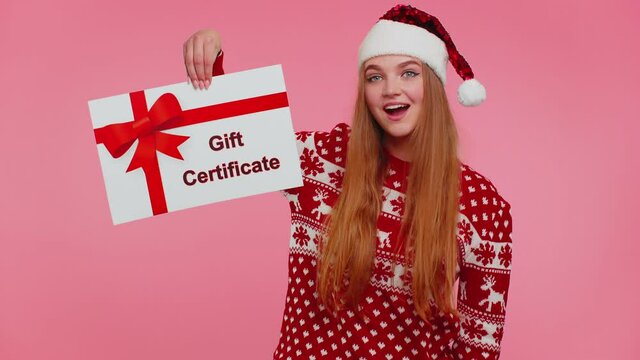 Adult 20s years old girl wears red New Year sweater and hat presenting card gift certificate coupon voucher isolated on pink studio wall background. Happy Christmas celebration merry holiday concept