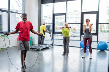 African american sportsman training with jump rope near sportswomen with dumbbells in gym.