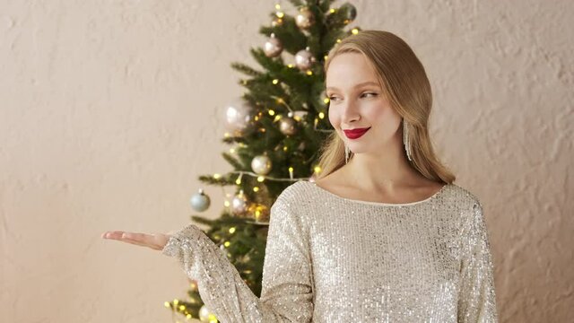 Young festive woman near christmas tree points with hand aside, shows copy space for your advertisement empty place for promotional image. Happy New Year promo mock up concept
