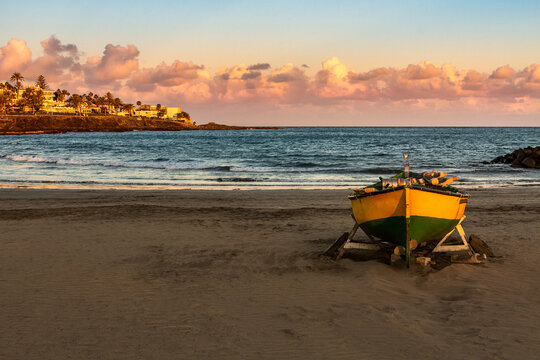 Colorful fisherman boat on a sandy beach shore. Sunset over coastline of atlantic ocean. El Jablillo beach in Costa Teguise, Lanzarote, Canary Islands, Spain. Travel destination seascape postcard.