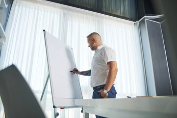 Side portrait of a middle aged Caucasian man in white casual shirt and tattooed arms writing business plan on flip chart