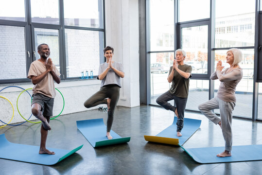 Interracial senior people standing in tree pose on yoga mats in sports center.