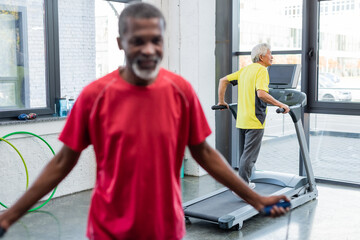 Senior asian man training on treadmill near blurred african american man with jump rope in gym.