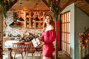beautiful young woman in red dress stands near Christmas tree in living room decorated for celebration of Christmas and New Year and opens gift box with wrapped Christmas presents