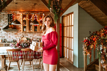 beautiful young woman in red dress stands near Christmas tree in living room decorated for celebration of Christmas and New Year and opens gift box with wrapped Christmas presents