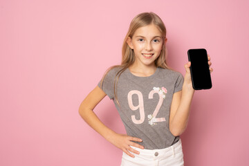 Smiling little girl in casual summer t-shirt showing mobile phone screen standing isolated over pink background.