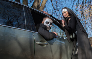 A man in the guise of a dead man in a car and a nun- witch near the hood of a car on Halloween.