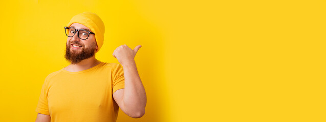 smiling man showing thumb up gesture and pointing at empty space  over yellow background, panoramic layout