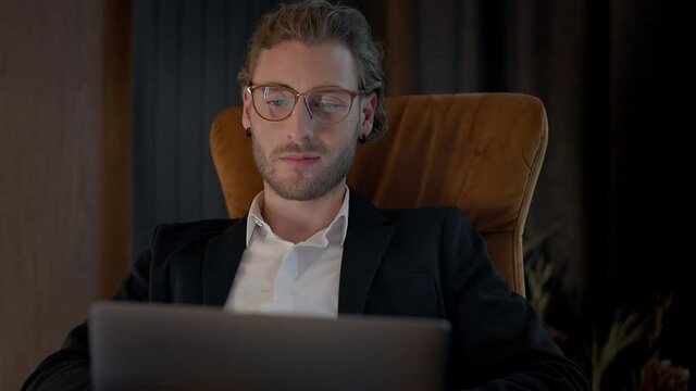 Young Man Sitting At Table With Laptop On His Lap. He Is In Hotel Room. Wonderful Interior. Handsome Man With Beard And Glasses.